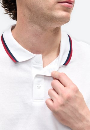 Nærbillede af en mand, der trækker i kraven på en hvid polo med røde og marineblå striber.