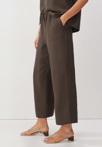 Pantalons larges marron à taille élastique, avec une texture lisse et des poches latérales, associés à des sandales minimalistes vert clair.
