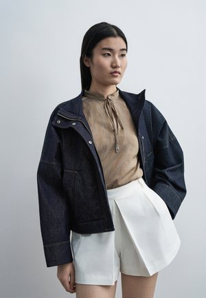 Jeune femme portant une veste en jean foncée, un chemisier beige à nouer au col et un short plissé blanc, debout devant un fond clair uni.
