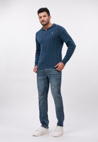 Bărbat îmbrăcat cu un pulover albastru din tricot, blugi albaștri închis decolorati și adidași albi, stând pe un fundal simplu.