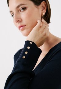 Marineblauer Pullover mit tiefem V-Ausschnitt und goldfarbenen Knopf-Details an den Ärmeln. Der Stoff wirkt weich und glatt und schafft eine figurbetonte Silhouette.