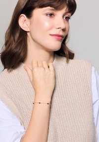 Złota bransoletka z czarnymi akcentami na smukłym łańcuszku, noszona na nadgarstku, w połączeniu z teksturowanym beżowym swetrem. Gładki i elegancki design.