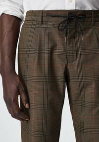 Brązowe spodnie w kratę z czarnymi i czerwonymi akcentami, elastyczny pas i zapięcie na sznurki. Materiał ma teksturowany wzór.