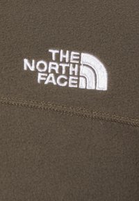 Olive zielony polarowy materiał o gładkiej teksturze; posiada biały haftowany logo "The North Face" przedstawiający design w postaci pół-łuku.