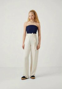 Top blu scuro senza spalline abbinato a pantaloni crema con vita alta e larga, fissati da una cintura annodata. La modella indossa scarpe con plateau nere. Tessuto morbido, vestibilità comoda.