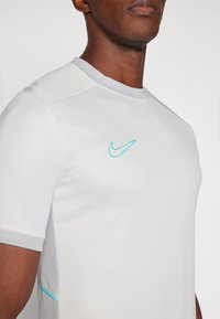 Bílý sportovní tričko s krátkými rukávy, s tyrkysovým logem Nike, síťovými bočními panely a kulatým výstřihem.