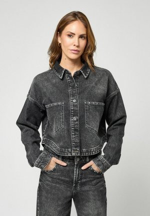 Frau trägt eine schwarze gewaschene Jeansjacke und passende hoch taillierte Jeans mit den Händen in den Taschen, sie steht vor einem schlichten hellen Hintergrund.