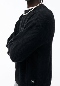 Pull noir tricoté avec une finition texturée, un col côtelé et des détails aux poignets. Présente une petite étiquette de marque près de l'ourlet.