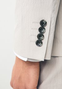 Nahaufnahme eines beigen Anzugsärmels mit vier dunklen Knöpfen und einem weißen Hemdärmelsaum, der über dem Handgelenk einer Person sichtbar ist.