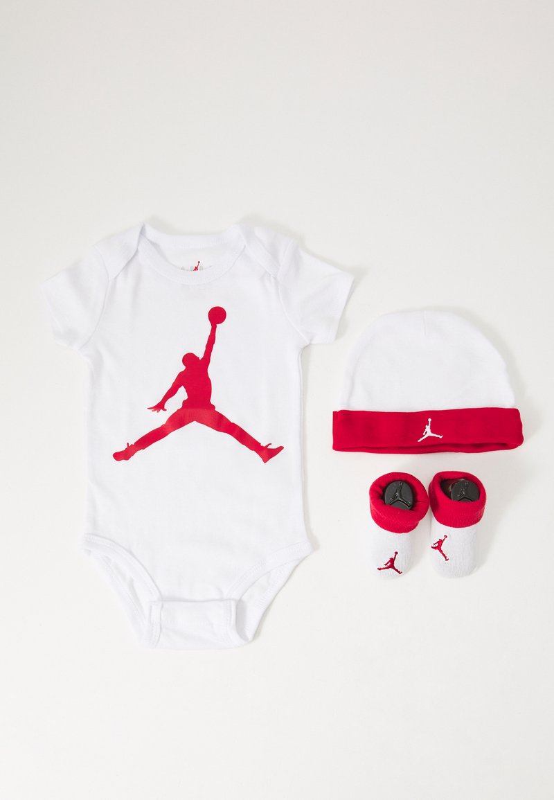 Un body blanc pour bébé avec un logo Jumpman rouge, accompagné d'un bonnet blanc à accents rouges et de chaussons assortis rouges et blancs.