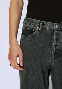 Lewa ręka z zielonym pierścionkiem spoczywa obok uda, ubrana w czarne zmywane dżinsy i czarną bluzkę na jasnym tle.