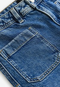 Los pantalones cortos de mezclilla en un lavado azul medio presentan un ajuste clásico, un diseño de cinco bolsillos y detalles de costura. Tejido texturizado con un ligero desgaste.