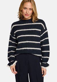 Femme portant un pull douillet bleu marine avec des rayures horizontales blanches et des poignets à volants, associé à un pantalon large bleu marine foncé.