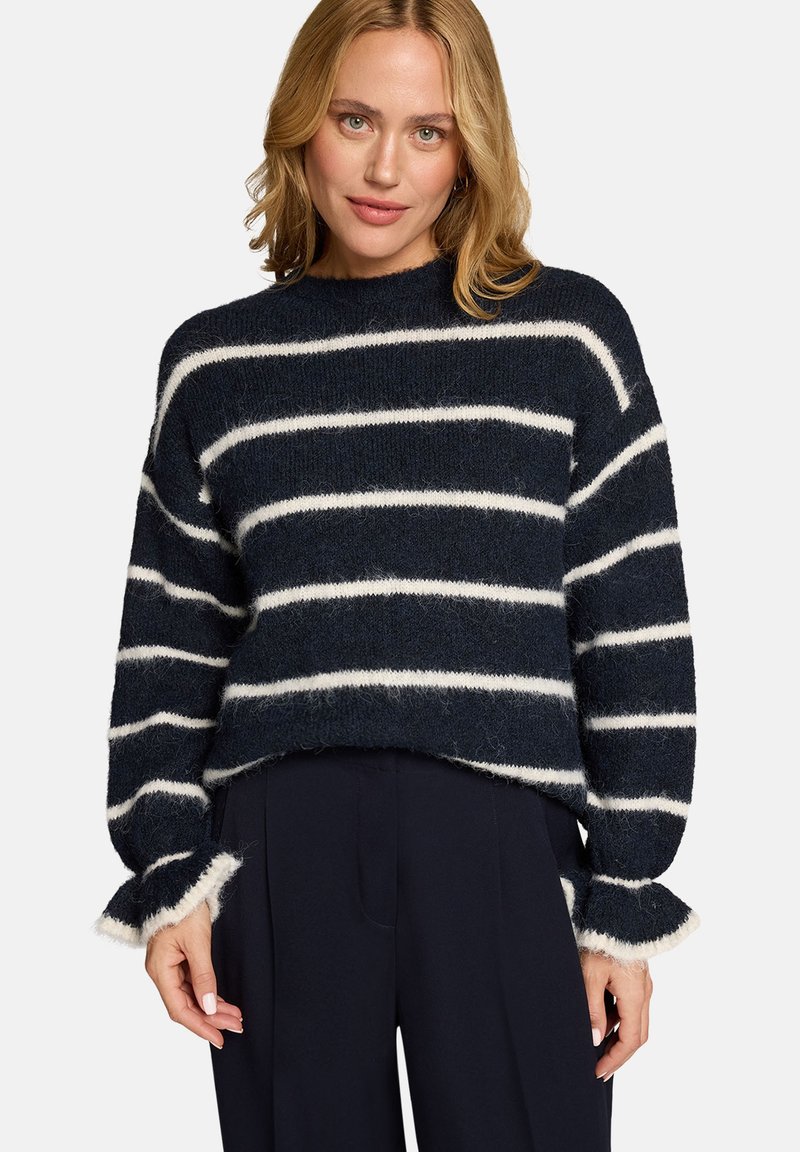 Femme portant un pull douillet bleu marine avec des rayures horizontales blanches et des poignets à volants, associé à un pantalon large bleu marine foncé.