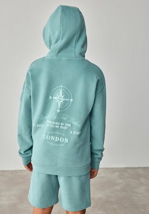 Kapuzensweatshirt in hellem Türkis mit weißen grafischen Details, einschließlich eines Kompassdesigns und Text, kombiniert mit passenden Shorts.