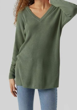 Model trägt einen langärmligen, lockeren V-Ausschnitt-Pullover in olivgrün, kombiniert mit blauen Jeans, und steht vor einem neutralen Hintergrund.