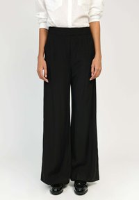 Pantalons larges noirs avec une texture lisse et une taille élastique, associés à une chemise blanche à boutons, sur un fond neutre.