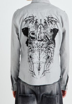 Rückseite eines gestreiften grauen Hemdes mit schwarzem Grafikdesign, das das Wort "SIN", abstrakte Flügel und symmetrische Muster zeigt, getragen von einer Person.