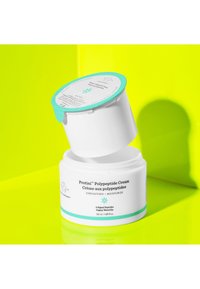 White jar with a turquoise lid; label details in black and turquoise. Cream texture visible inside, set against a bright neon yellow background.