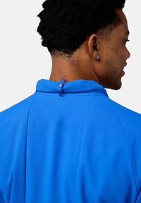 Giacca sportiva blu con colletto alto, tessuto texturizzato e un piccolo dettaglio ad anello sul retro. Nessun fermaglio o logo visibile.