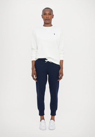 Femme debout portant un sweat-shirt blanc avec un petit logo, un pantalon de jogging bleu marine avec un cordon de serrage, et des baskets blanches sur un fond uni.