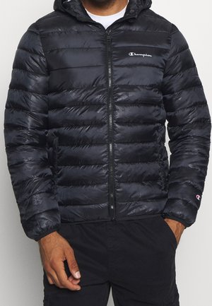 Hombre con chaqueta negra acolchada Champion con capucha y cremallera, camiseta blanca y pantalones negros, con las manos en los bolsillos.