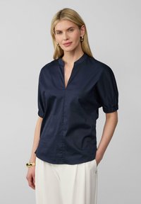 Blusa blu navy con colletto alla cinese, maniche a sbuffo corte con polsini arricciati e orlo dritto. Tessuto morbido e leggero.
