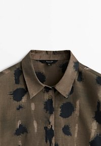 Ruskea paita, jossa on musta abstrakti kuvio, lyhyet kaulukset ja kaksi nappia. Valmistettu kevyestä kankaasta, jossa on pehmeä ja sileä rakenne.