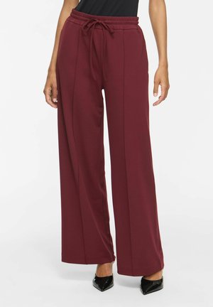 Femme portant un pantalon bordeaux taille haute à larges jambes avec une taille à cordon et des talons noirs à bout pointu, debout devant un fond uni.