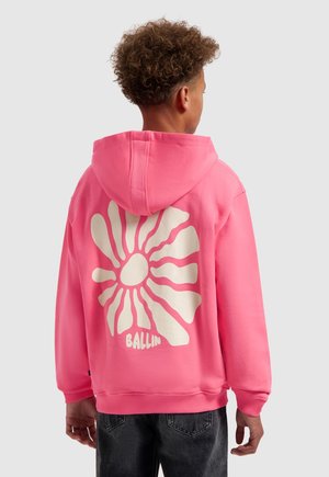 Jonge persoon met krullend haar draagt een felroze hoodie met een grote witte abstracte bloem en het woord "BALLIN" op de achterkant.