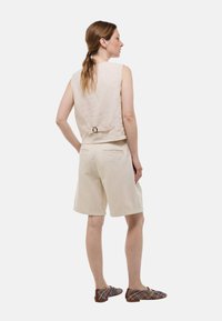 Top sans manches en lin beige avec un détail de boucle dans le dos, associé à un short beige et des chaussures à enfiler à motifs tissés.