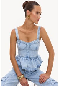 Femme aux cheveux attachés en chignon, portant un corset en denim bleu clair et un pantalon assorti, accessoirisée d’un bracelet et de boucles d’oreilles en or, assise les mains posées.