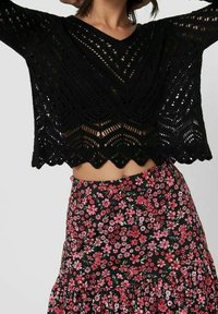 Schwarzer, kurzer Strickpullover mit gezacktem Saum und geometrischem Muster. Kombiniert mit einem schwarzen Blumenrock, der pinke Blumen und Rüschendetails aufweist.