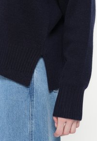 Maglione a maglia blu scuro con polsini e orlo a coste. Presenta spacchi laterali e un design rilassato, abbinato a jeans blu chiaro.