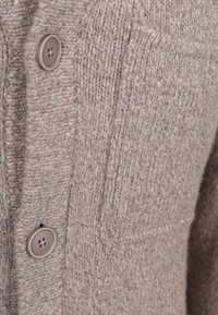 Gestructureerde beige cardigan van gebreide stof, met twee grote bruine knopen en een klein zakje aan de linkerkant.