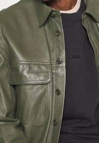 Olivgrön läderjacka med knäppning framtill, stor bröstficka och spetsig krage, lagd över en svart sweatshirts med logotypdetalj.