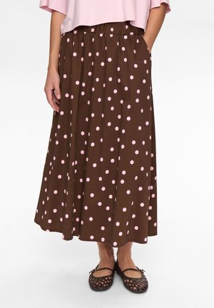 Brauner, bodenlanger Rock mit hohem Bund, versehen mit einem rosa gepunkteten Muster und Seitentaschen. Kombiniert mit braunen flachen Schuhen.