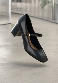 Chaussure Mary Jane en cuir verni noir avec un talon épais et une bride à boucle dorée, flottant au-dessus d'un sol gris clair.