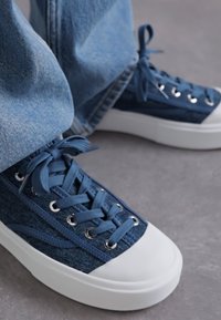 Denimblå sneakers med hvite gummisåler, som har metalløye, flate lisser og teksturert stoffdetaljering på sidene.