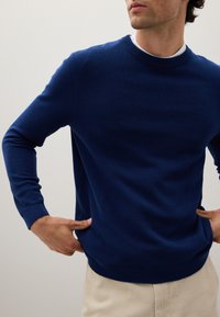 Maglione blu lavorato a maglia con superficie testurizzata, scollatura tonda e polsini e orlo a coste. Indossato sopra una camicia bianca con pantaloni beige.