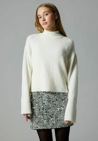 Kudum valge sviiter kõrge kaeluse ja pikkade varrukatega, kombineeritud sädelevate mustade ja hõbedaste mini-seelikutega. Sile tekstuur, lühendatud disain.