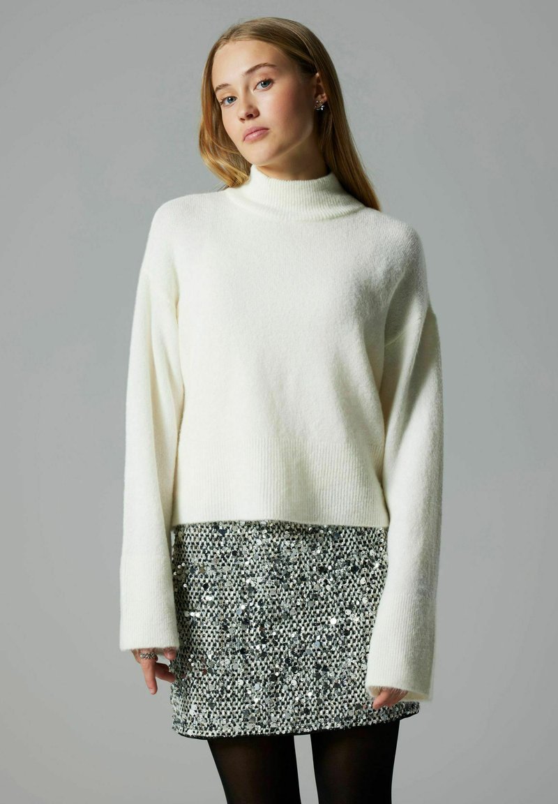 Kudum valge sviiter kõrge kaeluse ja pikkade varrukatega, kombineeritud sädelevate mustade ja hõbedaste mini-seelikutega. Sile tekstuur, lühendatud disain.