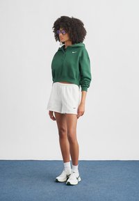 Sweat à capuche court vert avec logo Nike, associé à un short blanc. Le mannequin porte des lunettes de soleil violettes et des chaussures de sport blanches avec des accents verts.