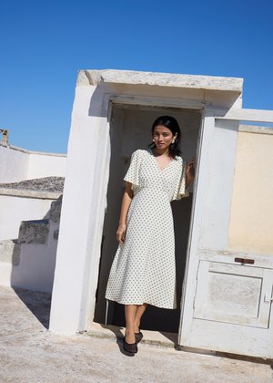 Kobieta w białej sukience midi w groszki i czarnych butach stoi w białym framudze drzwi pod czystym błękitnym niebem.