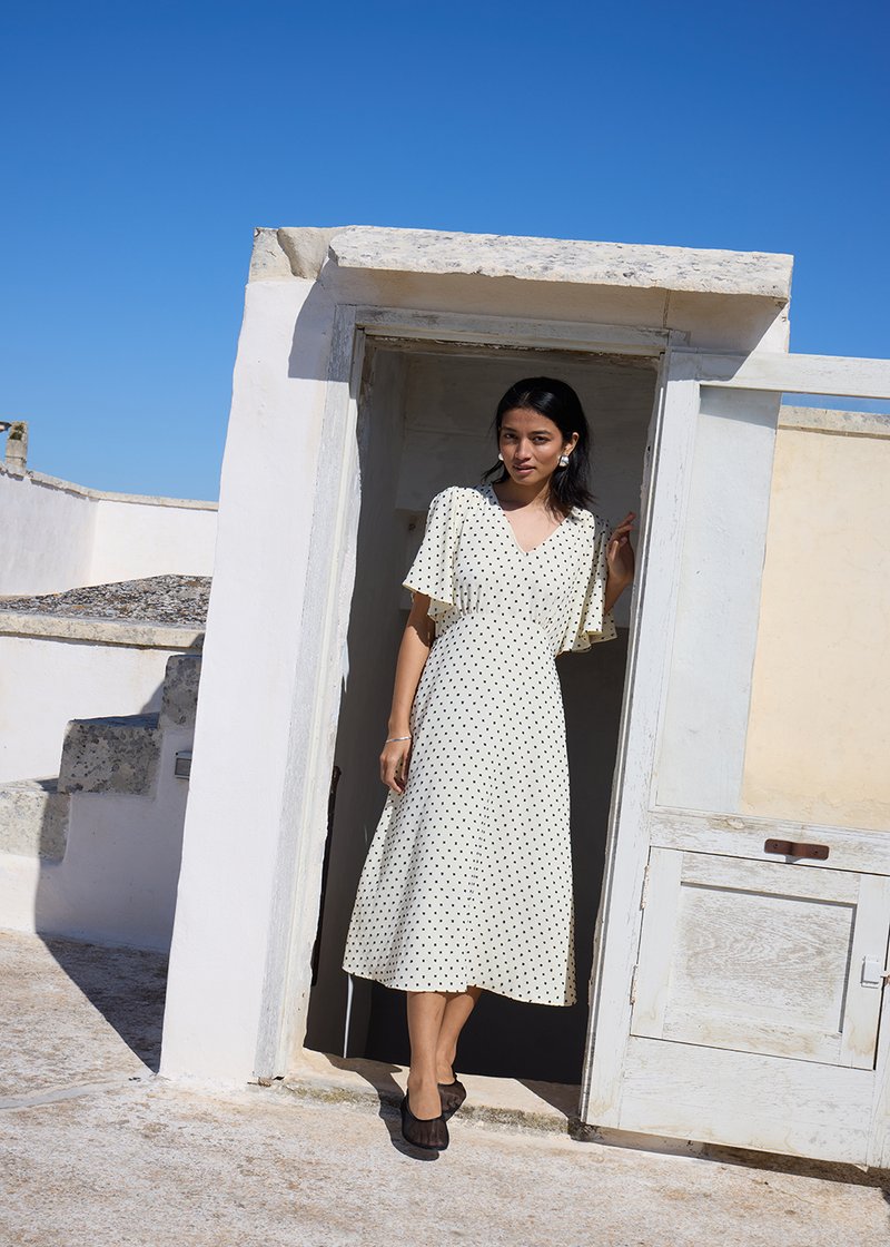 Donna con abito midi bianco a pois e scarpe nere, in piedi in uno stipite bianco sotto un cielo azzurro limpido.