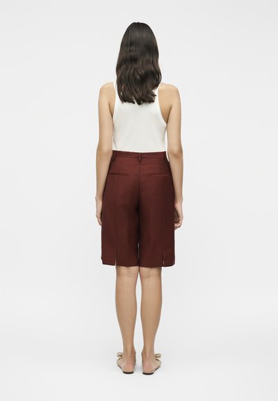 Γυναίκα με σκούρα κυματιστά μαλλιά που φοράει λευκό αμάνικο τοπ, σκούρες καφέ βερμούδες μέχρι το γόνατο και επίπεδα σανδάλια, όρθια με την πλάτη στραμμένη μπροστά.