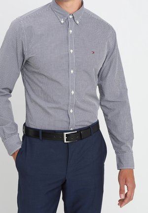Homme portant une chemise à carreaux bleu et blanc boutonnée, rentrée dans un pantalon bleu marine avec une ceinture noire, debout avec une main dans la poche.