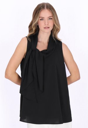 Tânără cu păr blond ondulat poartă un top negru fără mâneci, cu un detaliu de fundă drapată, stând în fața unui fundal alb.