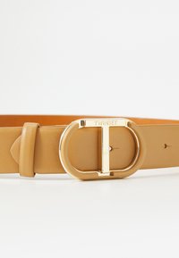 Tan leather belt featuring a gold-tone circular buckle with a "T" design. Smooth texture with a single loop and several adjustment holes.