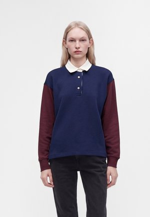 Jeune femme aux longs cheveux blonds porte une chemise à manches longues bleu marine et bordeaux avec un col blanc et un pantalon noir, debout devant un fond uni.